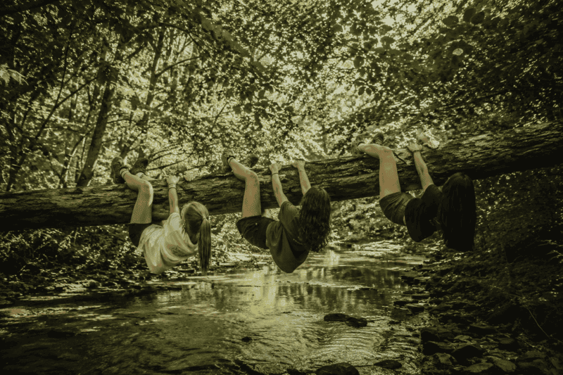 O que fazer com as amigas nas férias dentro e fora de casa 2 O que fazer com as amigas nas férias em uma floresta verde com três meninas brincando em um tronco sobre um riacho.